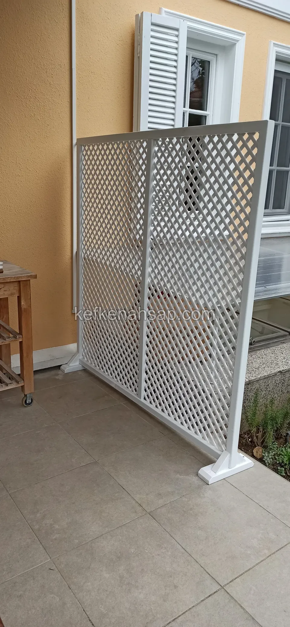 Sarı cepheli evin terasında, panjurlu pencerenin yanında beyaz boyalı çaprazlı ayaklı kafes panel; karo zeminde