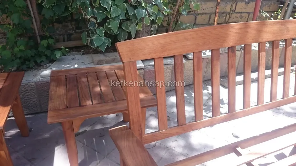 A reddish-varnished timber bench with an upright slatted back and a small slatted table in front of an ivy wall
