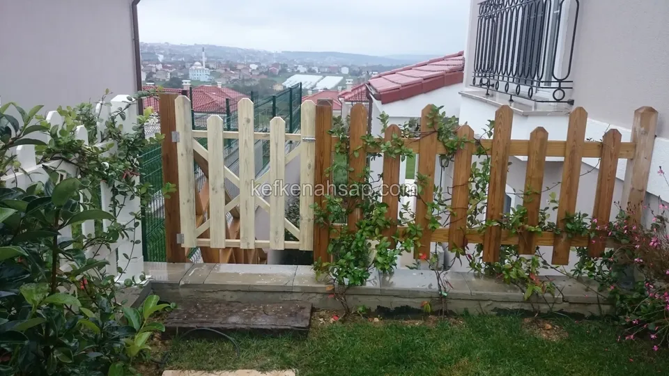 Sarmaşık sarılmış kahverengi çıtalı çit ve açık tonlu çıtalı kapı, arkada kiremit çatılı evler ve şehir manzarası