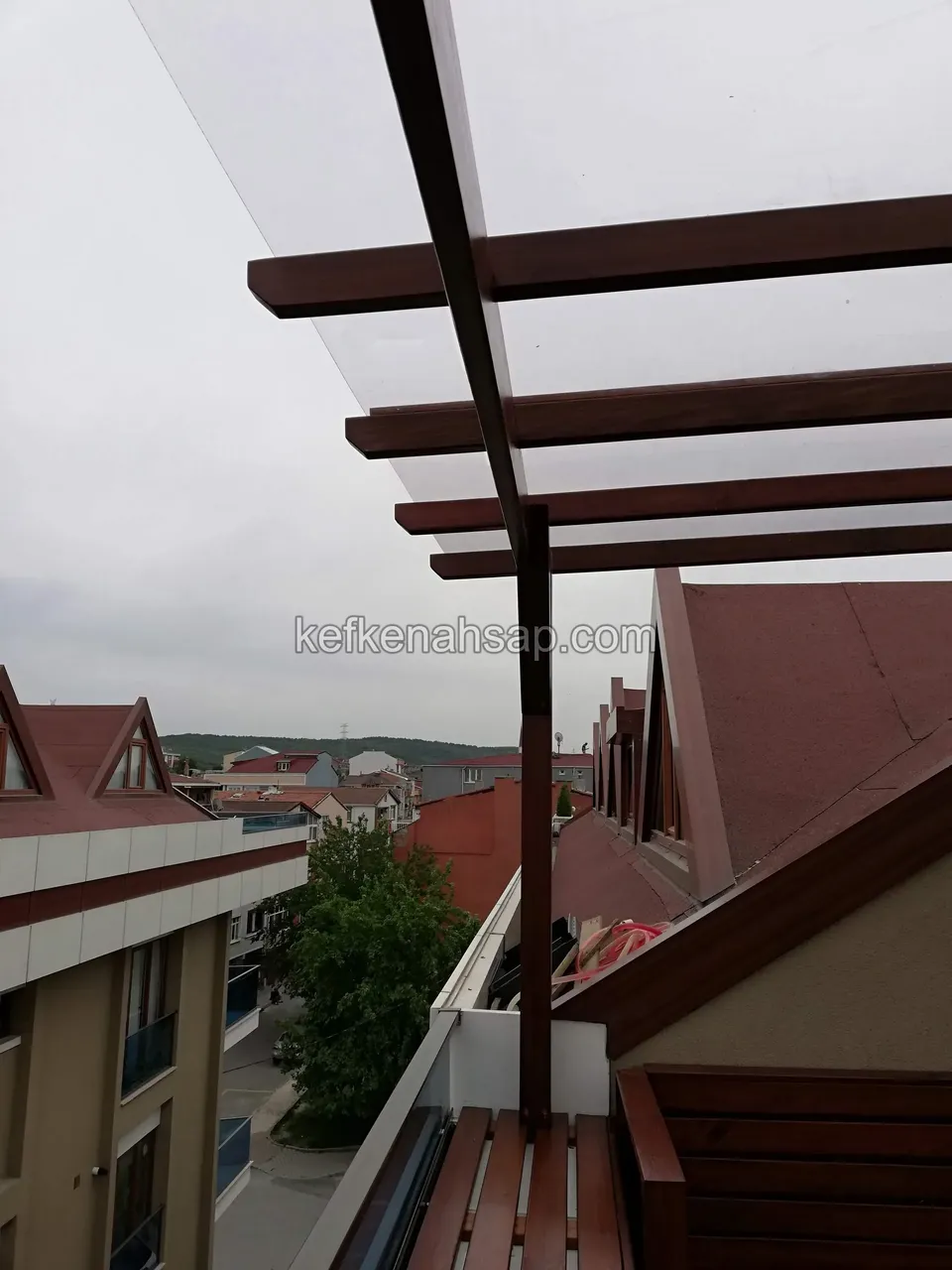 Şeffaf çatı örtüsü altında sıralanan ahşap pergola kirişleri, yanda ahşap bank ve çevrede çatılar