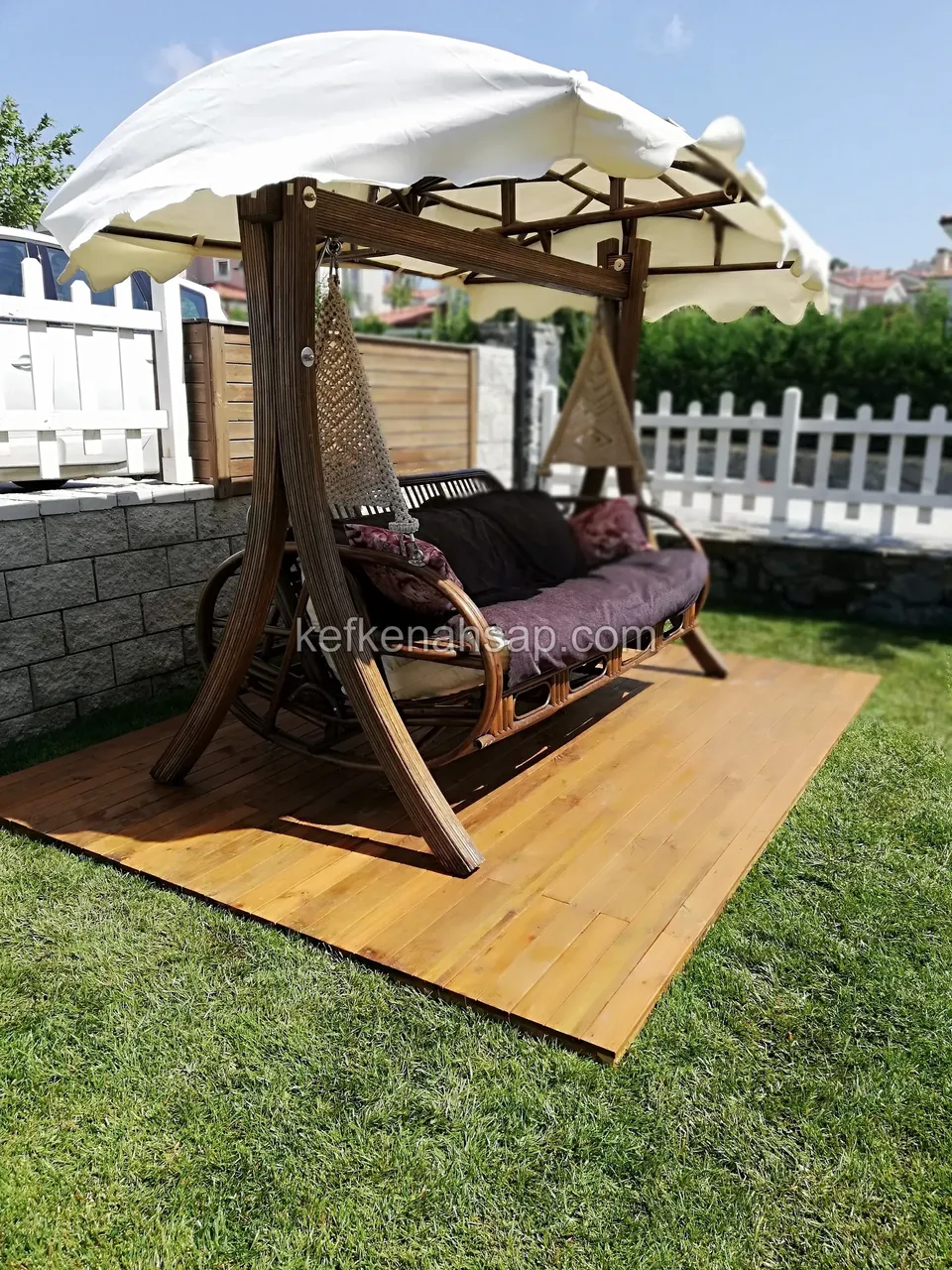 Beyaz çitin önünde, ahşap deck üstünde duran şemsiye tenteli ve minderli ahşap salıncak