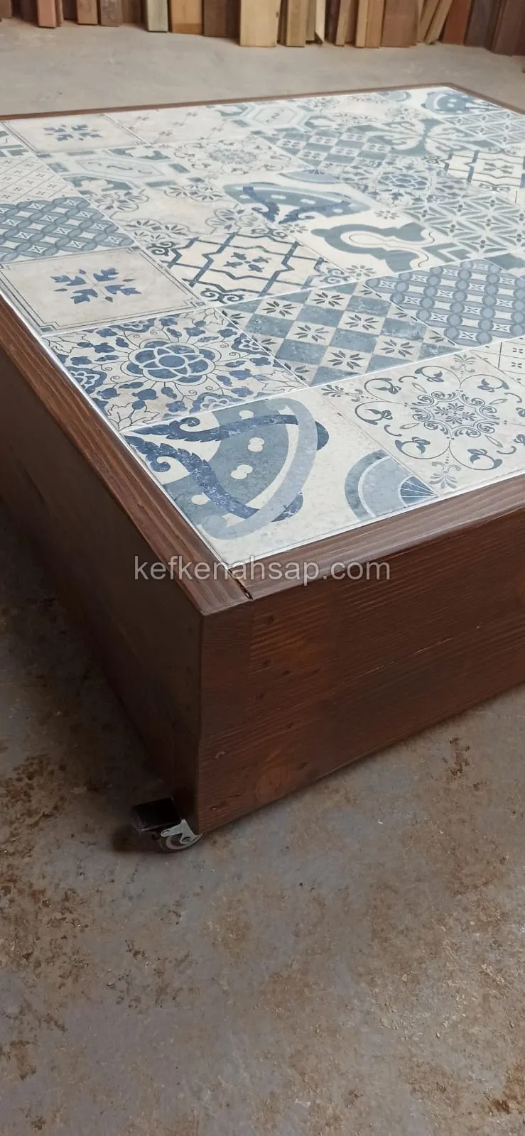 Low dark brown wooden table with a blue and white patterned ceramic tile top and casters, standing on a workshop floor