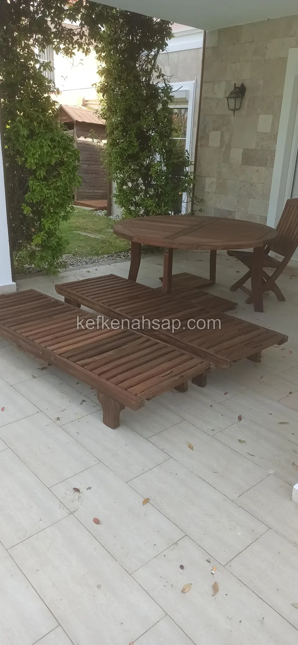 Auf der hellen Keramikterrasse am Hauseingang stehen zwei dunkel lackierte Holzliegen, ein runder Tisch und ein Klappstuhl