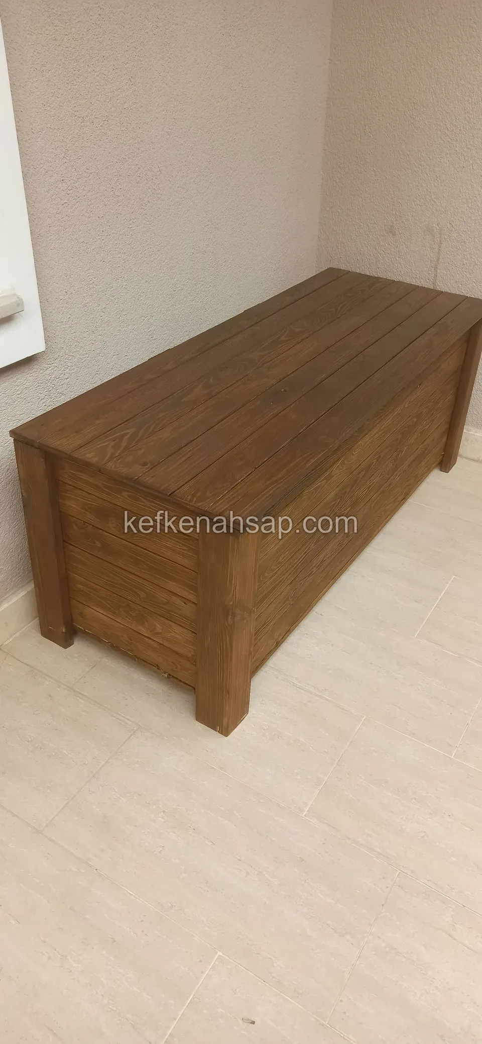 Eine braun lackierte Truhenbank aus Holzlatten mit Deckel in einer Ecke auf hellem Keramikboden