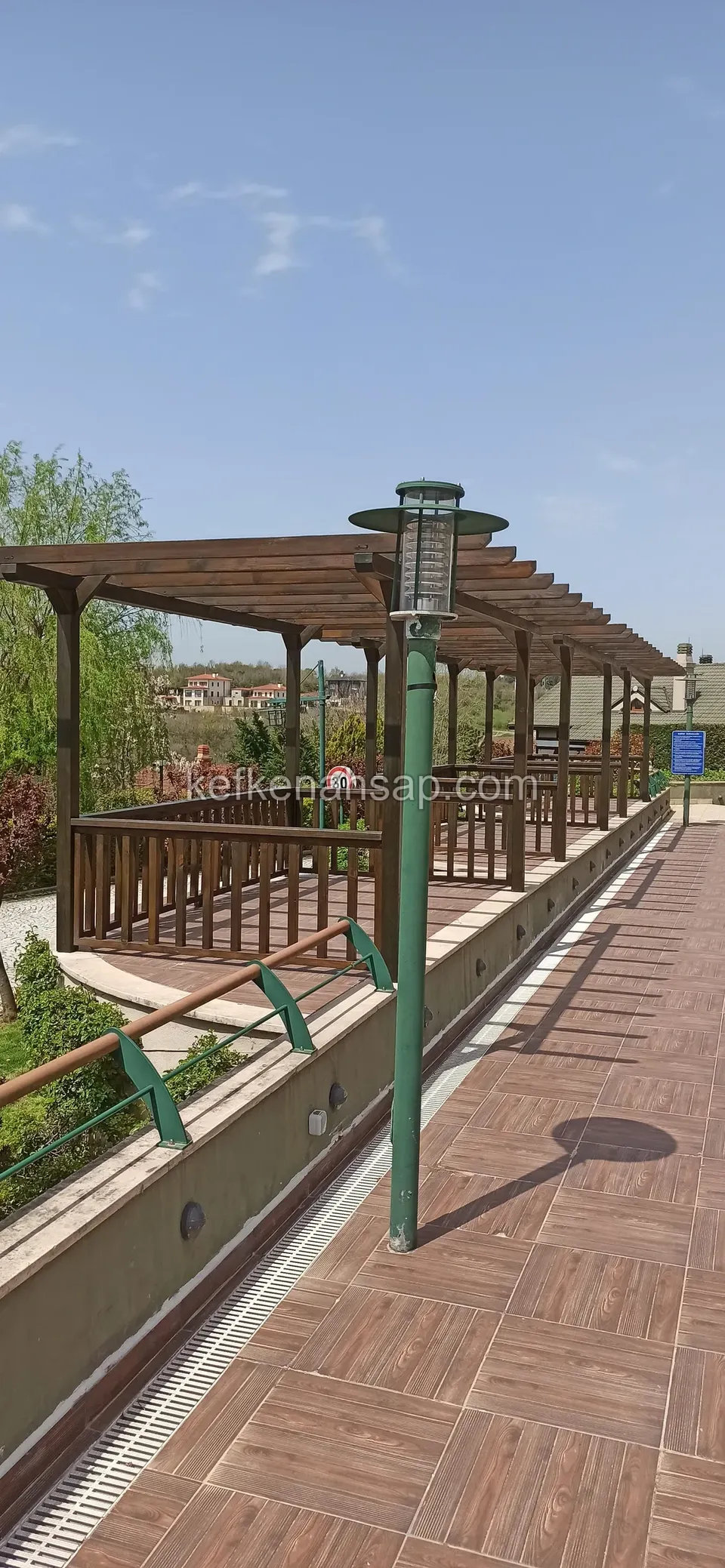 Ahşap görünümlü seramik zeminli yürüyüş yolunun yanında korkuluklu ahşap pergola ve yeşil lamba direği