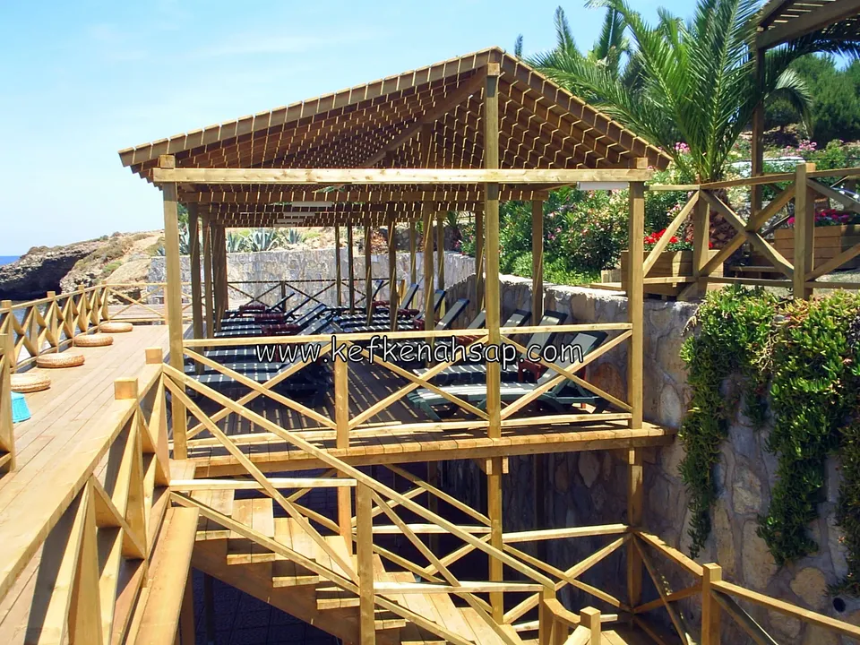 Erhöhtes Deck mit Liegen vor einer Steinmauer, darüber eine Pergola mit Tonnendach und eine Holztreppe