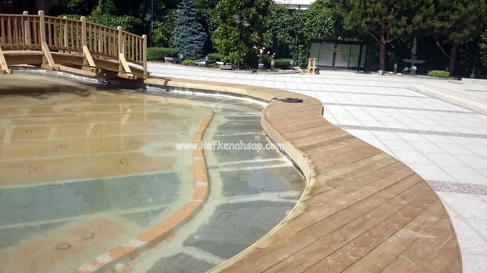 Sığ su dolu kavisli havuzun etrafındaki geniş deck kenarı ve havuzun üstündeki küçük korkuluklu ahşap köprü
