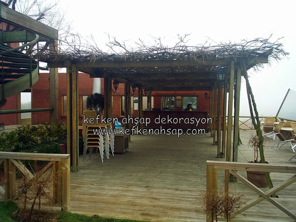 Sisli havada geniş ahşap deck üzerinde direkler, üzeri dallarla örtülü pergola, arkada tuğla bina ve spiral merdiven