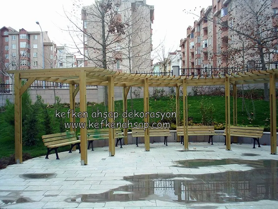 Apartmanların önündeki taş döşeli alanda, altında ahşap bankların sıralandığı açık renkli ahşap pergola