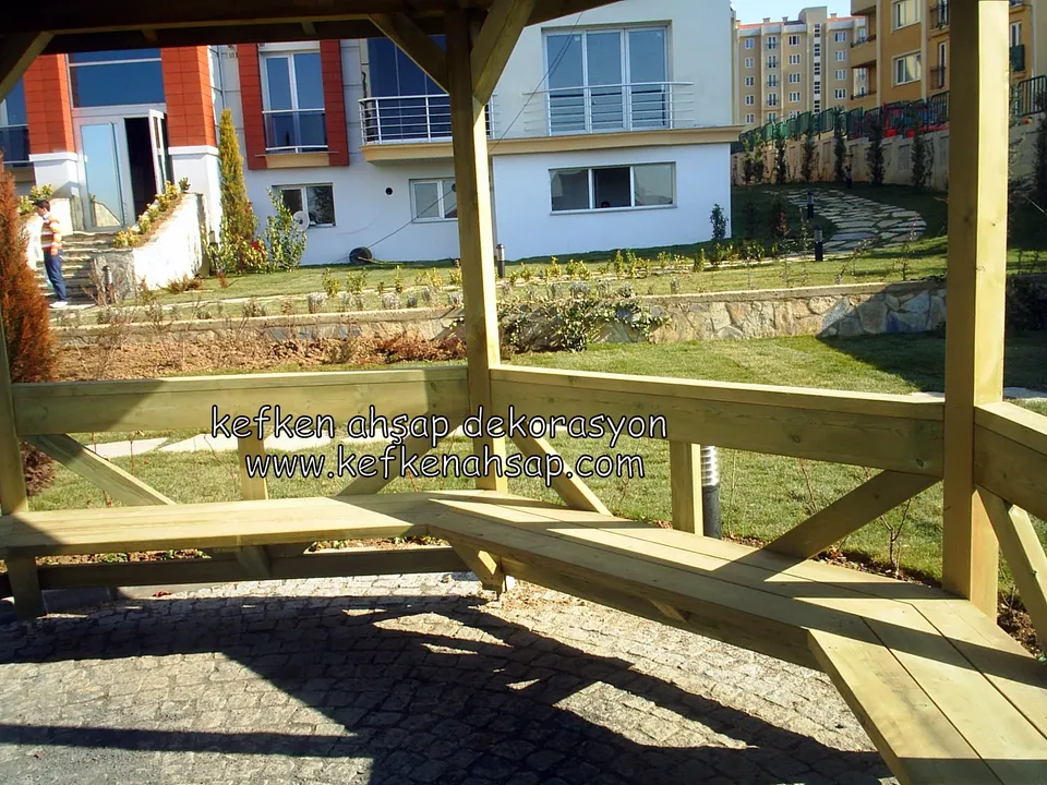 L-shaped bench under a light wooden pergola in a housing complex garden, with lawn, stone path and buildings behind