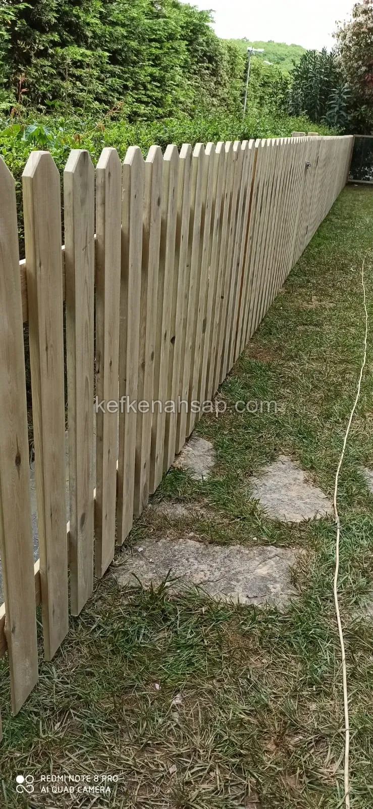 Çimenli bahçede sık dizilmiş, sivri uçlu açık renk ahşap çıtalardan yapılmış uzun çit; yanında taş basamaklar var