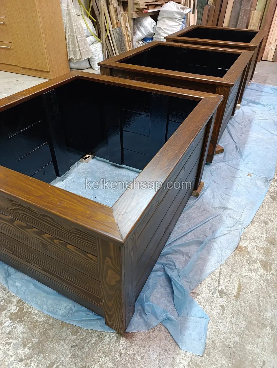 Square timber planters with black liners lined up on blue plastic, the front one seen into the inside