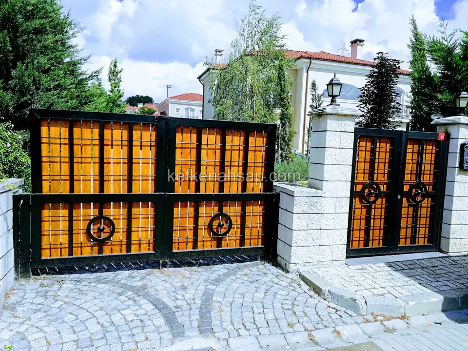 Küp taş döşeli girişte, turuncu-kahve ahşap dolgulu siyah çerçeveli geniş sürgülü kapı ve yanında daha dar yaya kapısı