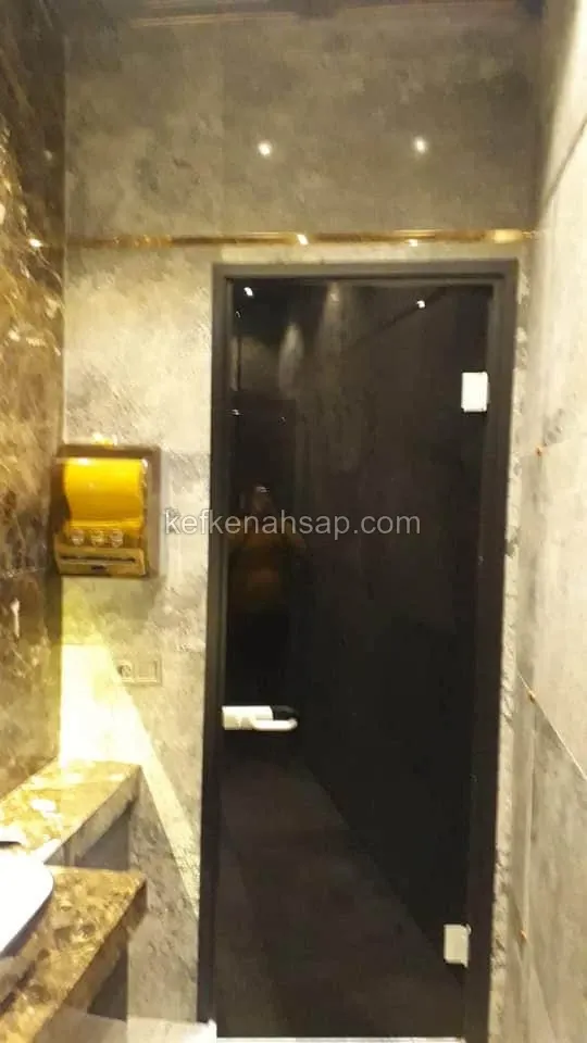 Gri sıvalı duvar ile kahverengi mermer tezgâh arasında siyah çerçeveli, füme camlı kapı