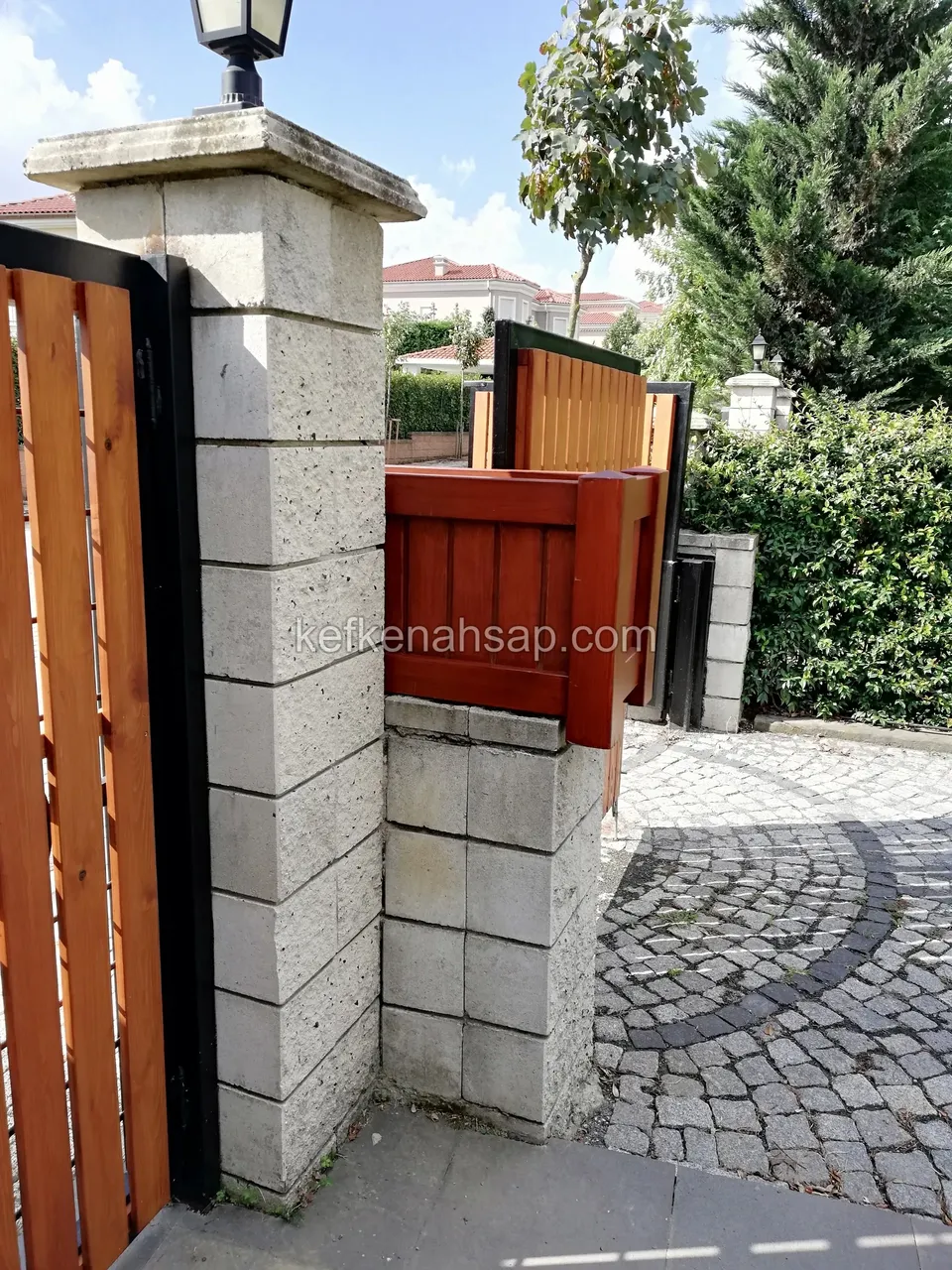 Beyaz taş sütunun yanında siyah çerçeveli, turuncu-kahve dikey tahtalı kapı ve sütun üstündeki kırmızı-kahve ahşap kutu
