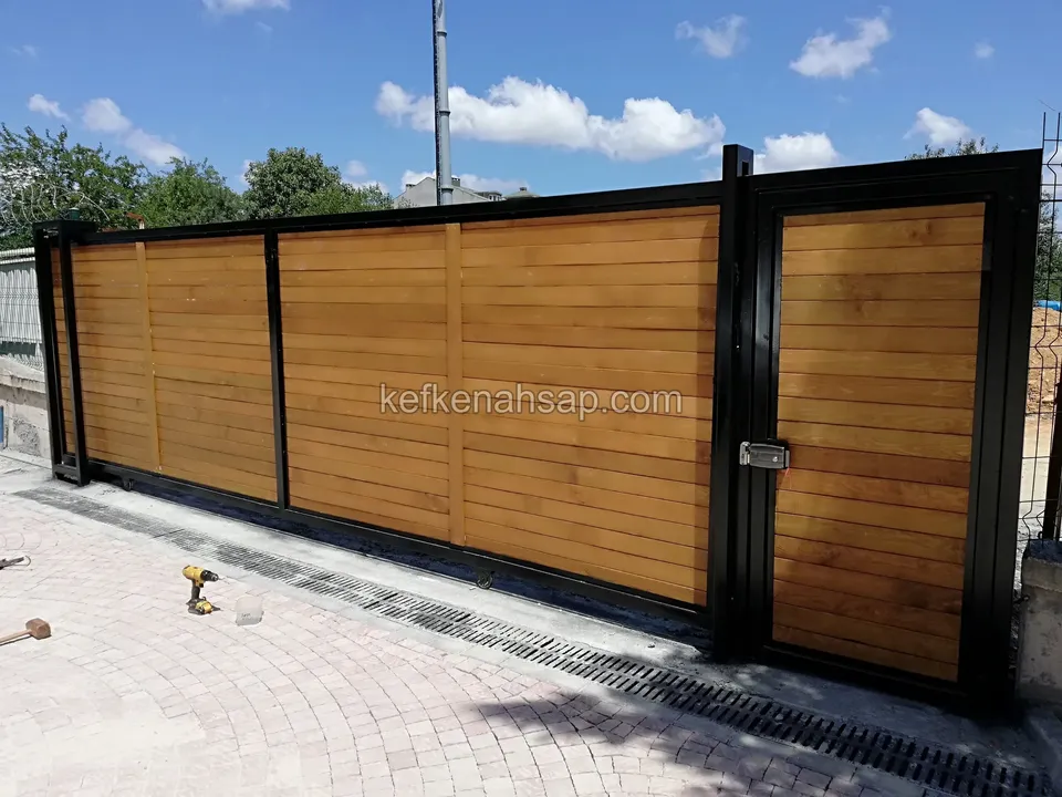 Parke taşlı girişte, siyah metal çerçeveli yatay tahta dolgulu uzun sürgülü kapı ve sağda yaya kapısı