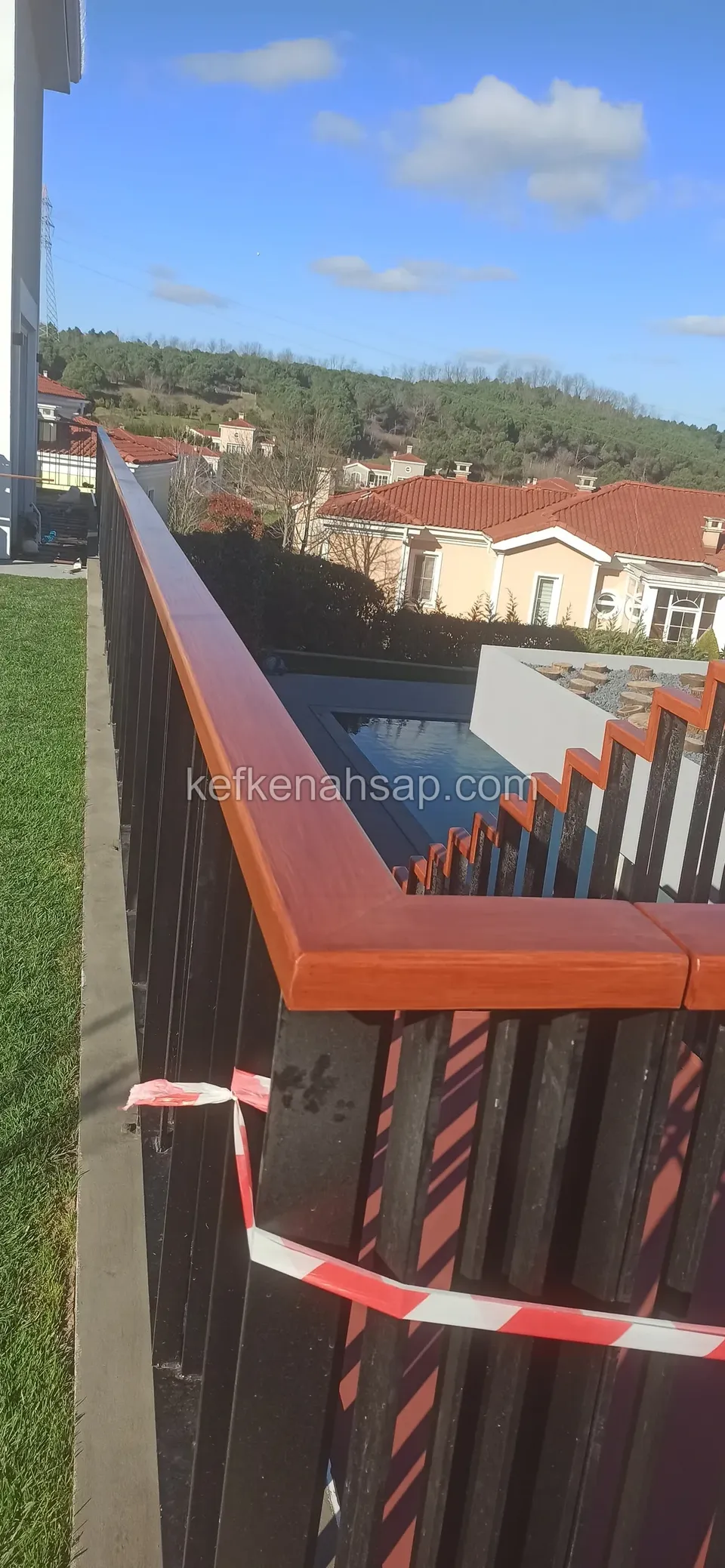 Çimlik kenarında siyah çıtalı korkuluğun üstünde geniş, turuncu tonlu ahşap küpeşte, çıtalara bant şeridi bağlı