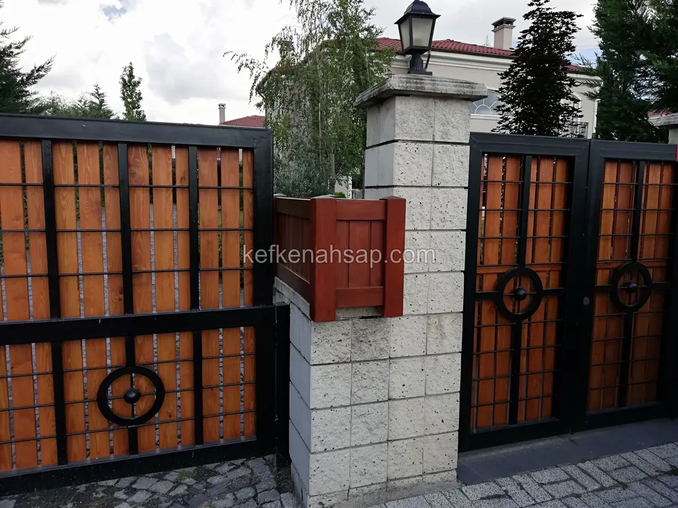 Taş sütunun iki yanında siyah çerçeveli, turuncu-kahve ahşap dolgulu bahçe kapıları; dairesel demir motifler