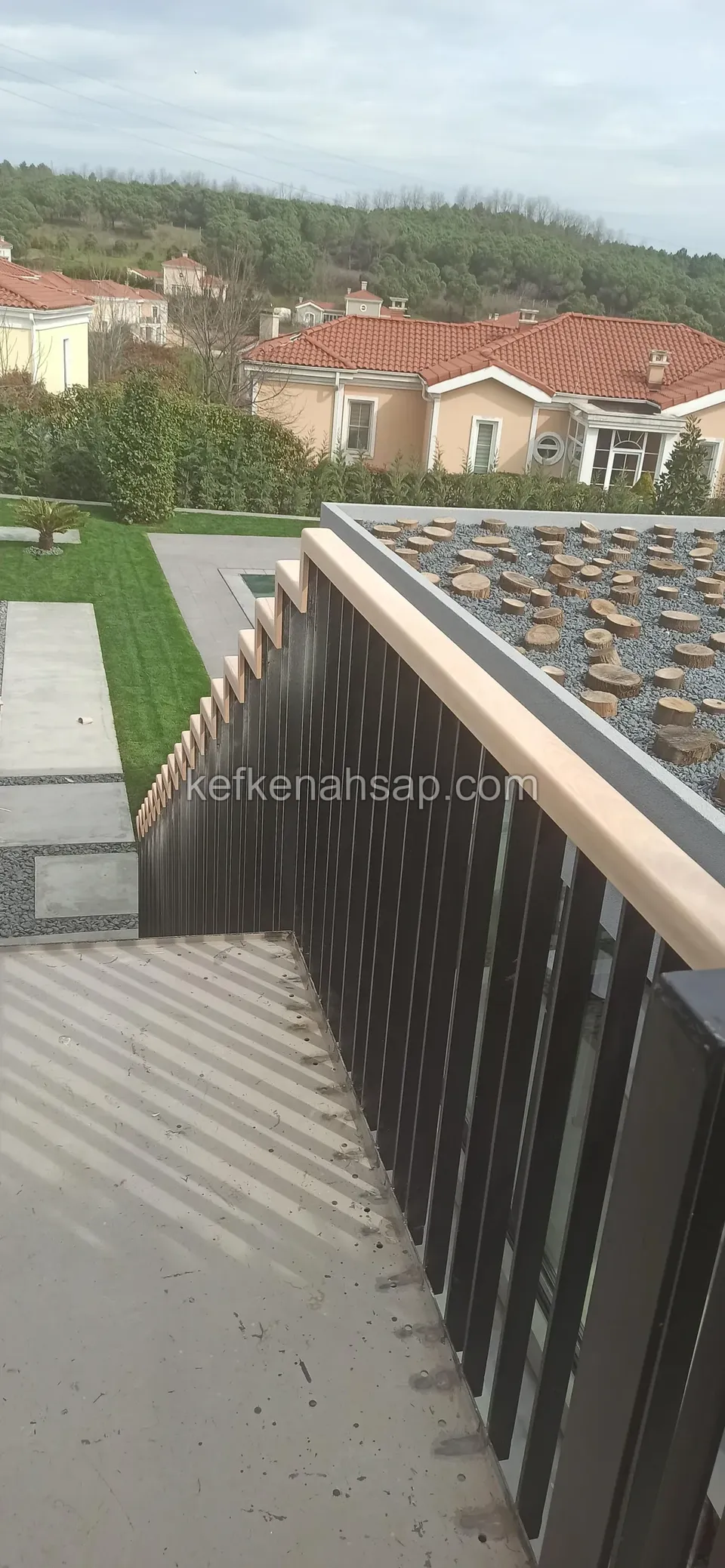 Merdiven kenarında siyah boyalı dikey çıtalı korkuluk, üstünde turuncu tonlu ahşap küpeşte, arkada konutlar
