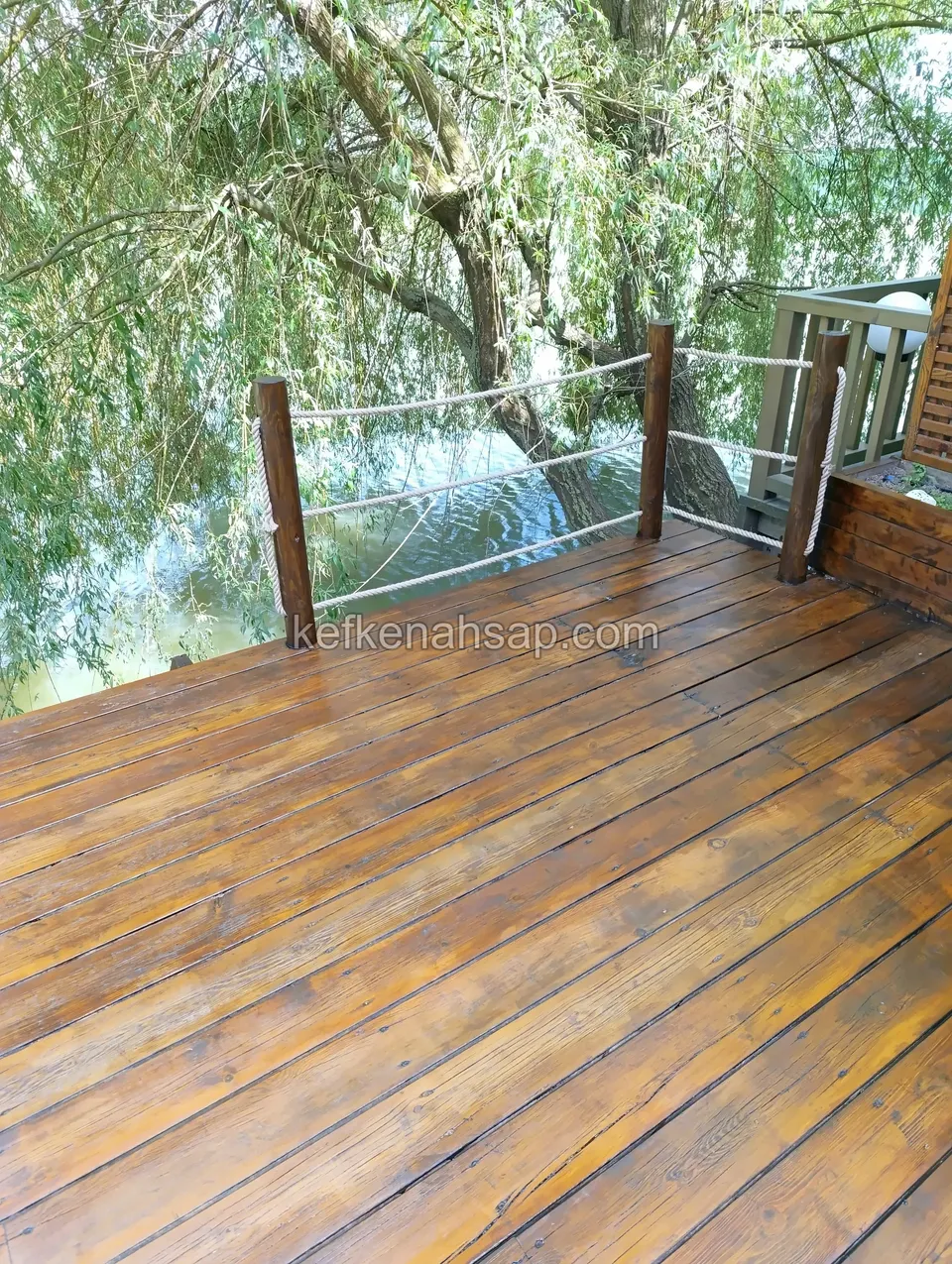 Polished deck facing a lake beneath a willow with hanging branches, with a timber railing strung with rope