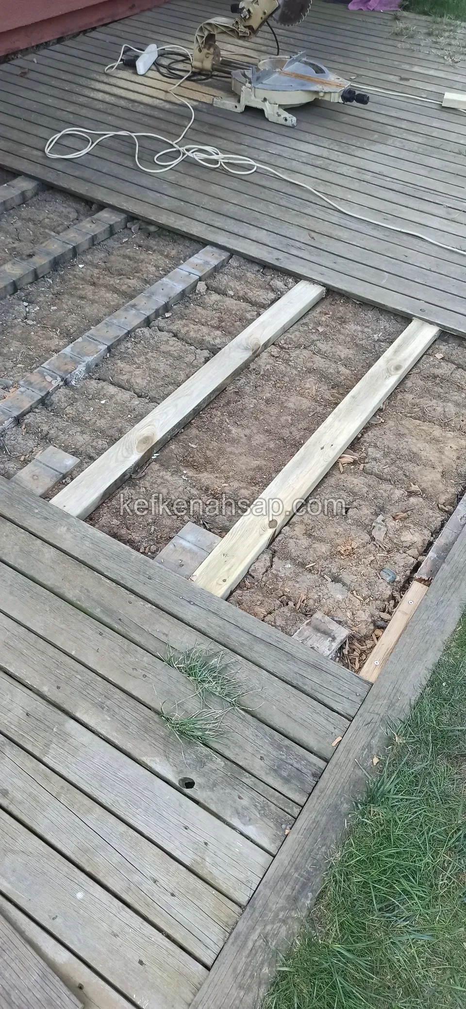 Eski gri deck tahtaları sökülmüş, kirişlerin arasında toprak ve gönye testere ile kablo görünüyor