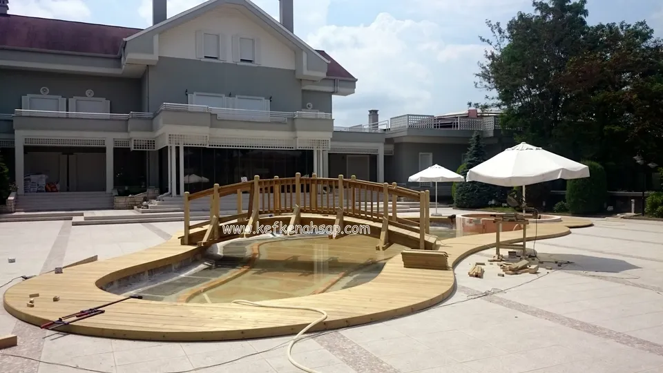 Su dolu havuzun çevresinde kıvrımlı ahşap deck ve ortasında korkuluklu kemerli köprü; arkada iki katlı bina ve şemsiyeler