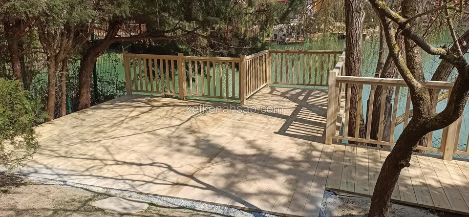 Broad pale deck among trees at the waterside, with a railing at the far end
