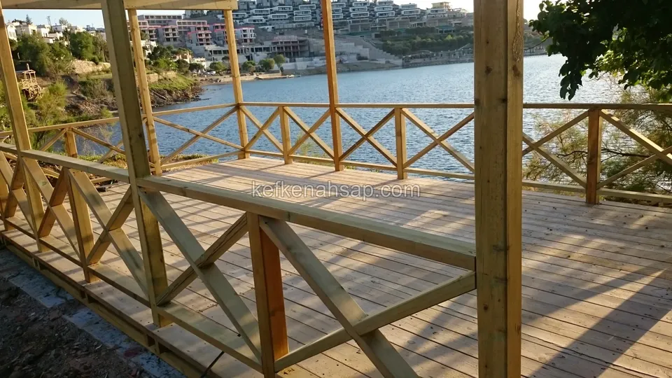 Suya bakan, çapraz korkuluklu ve pergola dikmeli ahşap deck; karşı kıyıda yapılar görünüyor
