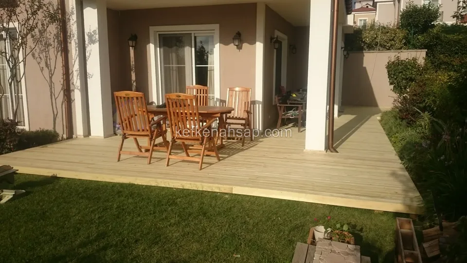 Zemin kat sundurmasının altından çime uzanan açık renkli deck, üzerinde ahşap masa ve dört sandalye