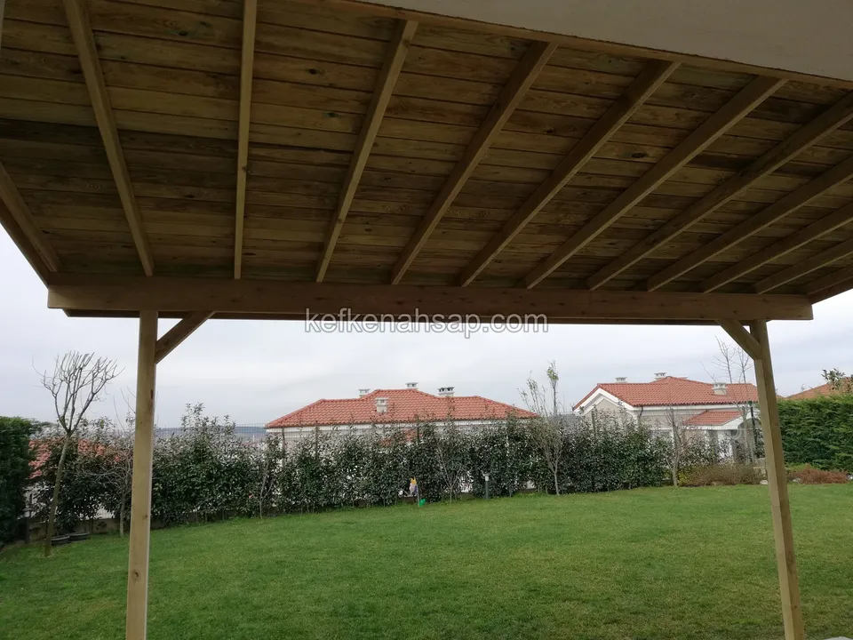 Sundurmanın altından görünen tahta kaplama ve kirişler; ileride çim bahçe ve komşu evler