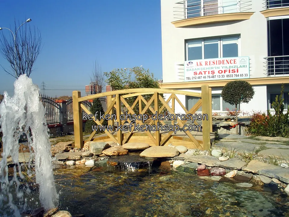 Taş kenarlı süs havuzunun üzerinde çapraz korkuluklu, hafif kemerli ahşap köprü; solda fıskiye, arkada apartman var.