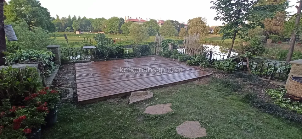 Тёмный красно-коричневый настил на газоне в саду у воды, со ступенчатыми плитами и цветами сбоку