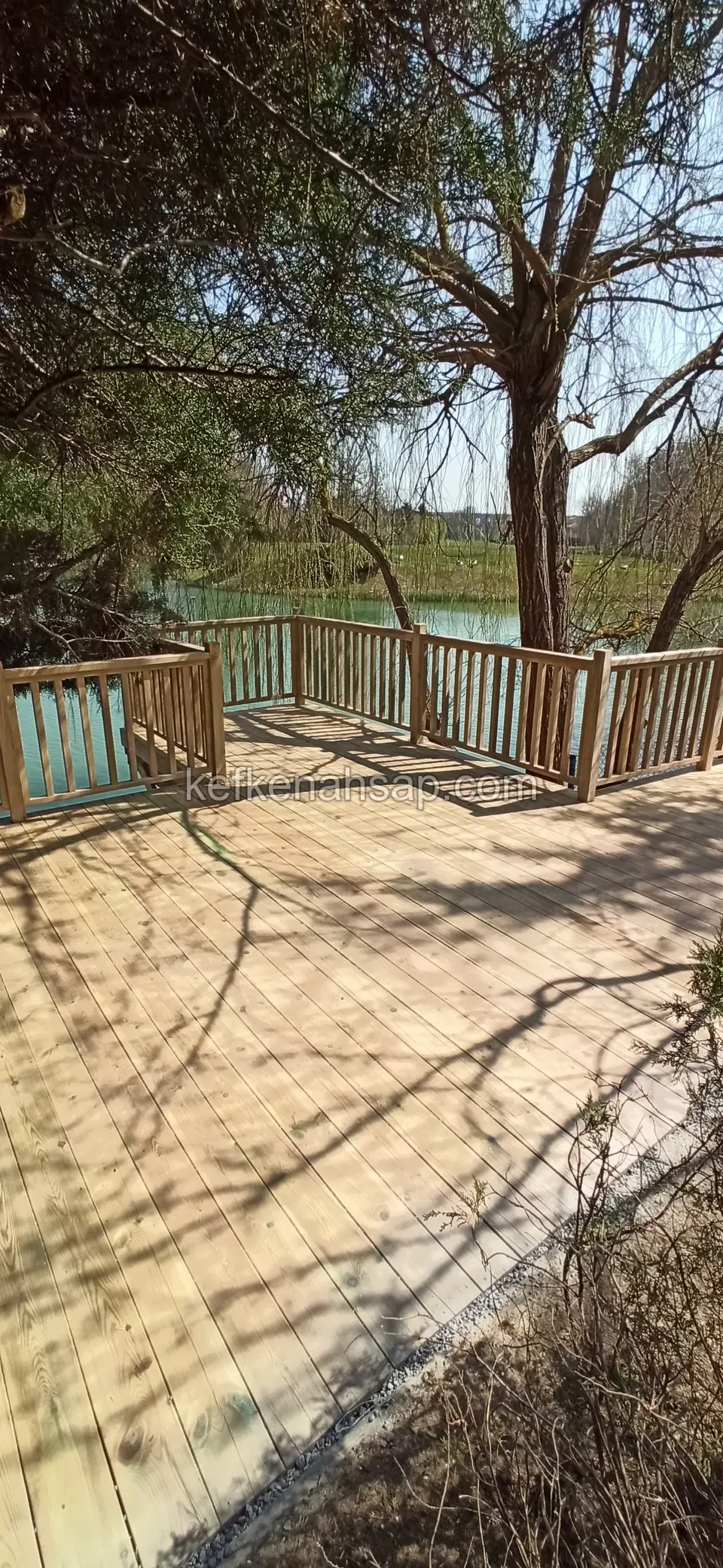 Suya bakan açık renkli deck ve üzerine düşen ağaç gölgeleri, uçta ahşap korkuluk