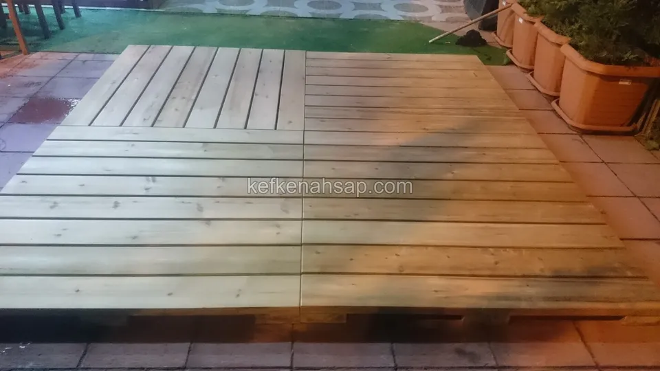 Parke taşlı zemine yerleştirilmiş açık renkli emprenyeli çam deck paneli, tahtalar iki farklı yönde, kenarda saksılar