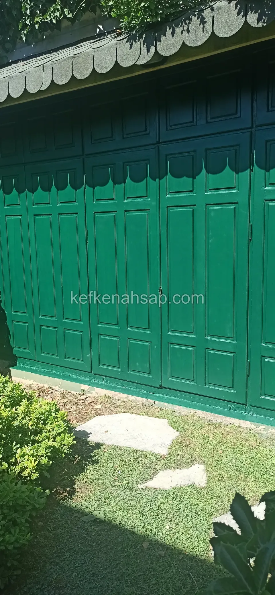 Çimenin içindeki taş basamaklı patikanın kenarında yeşil boyalı kasetli kapı yüzeyi ve üstünde açık renkli saçak