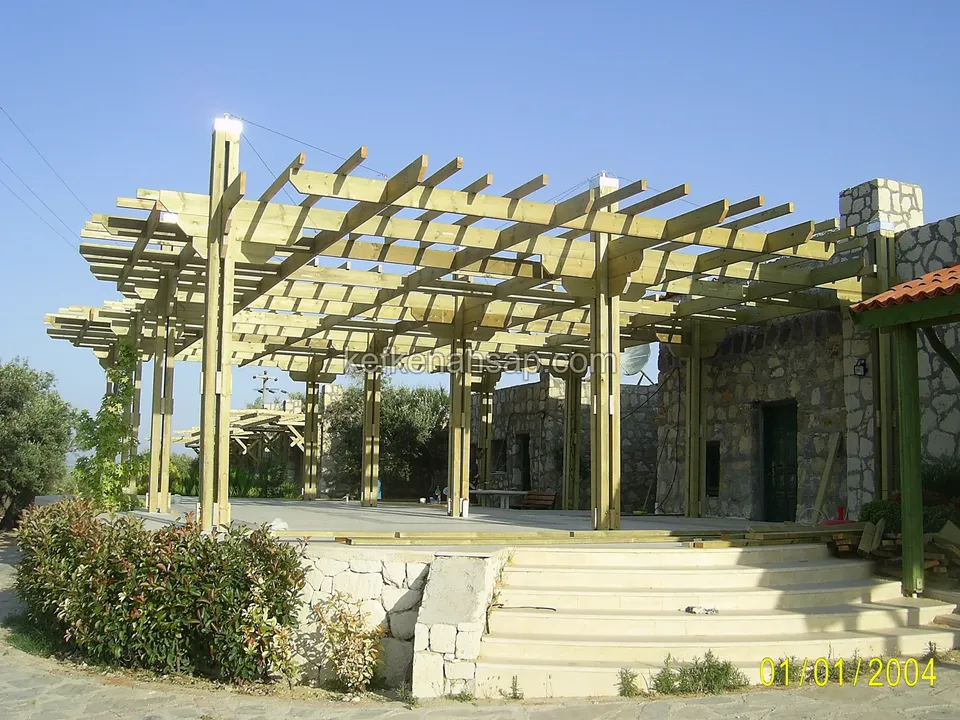 Taş kaplı binanın önünde yükseltilmiş zeminde geniş, açık renk ahşap pergola; önünde geniş basamaklar