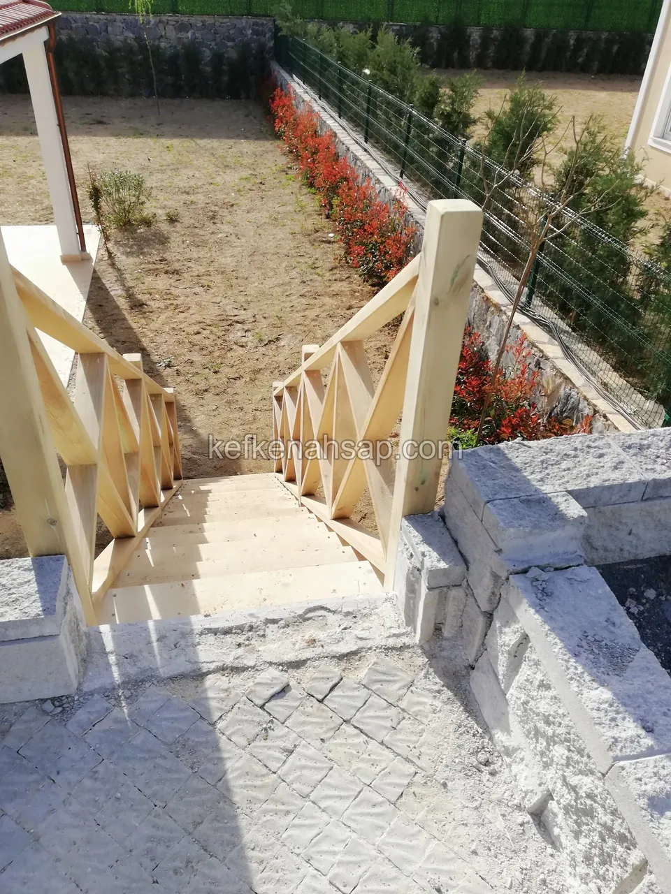 Holztreppe, die zwischen Steinblöcken hinabführt, rechts rotblättrige Sträucher und ein Maschenzaun