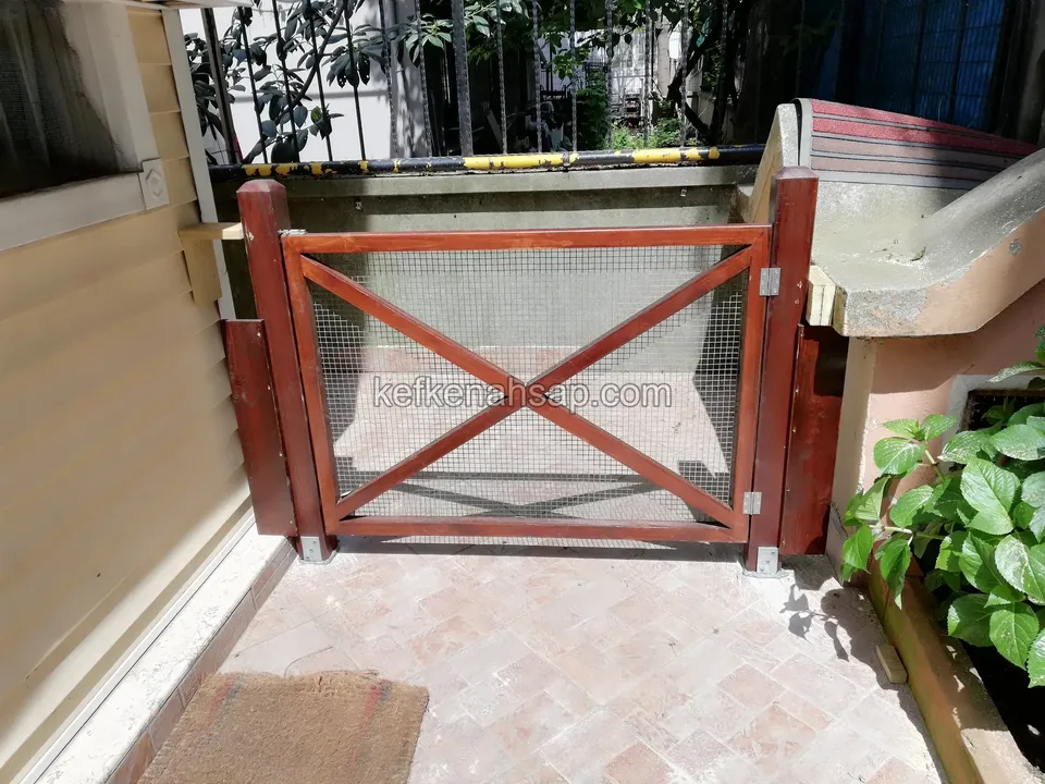 Taş döşeli dar geçitte, X çapraz kirişli ve tel örgü dolgulu kırmızı boyalı alçak ahşap kapı; yanda bitkili saksı ve duvar