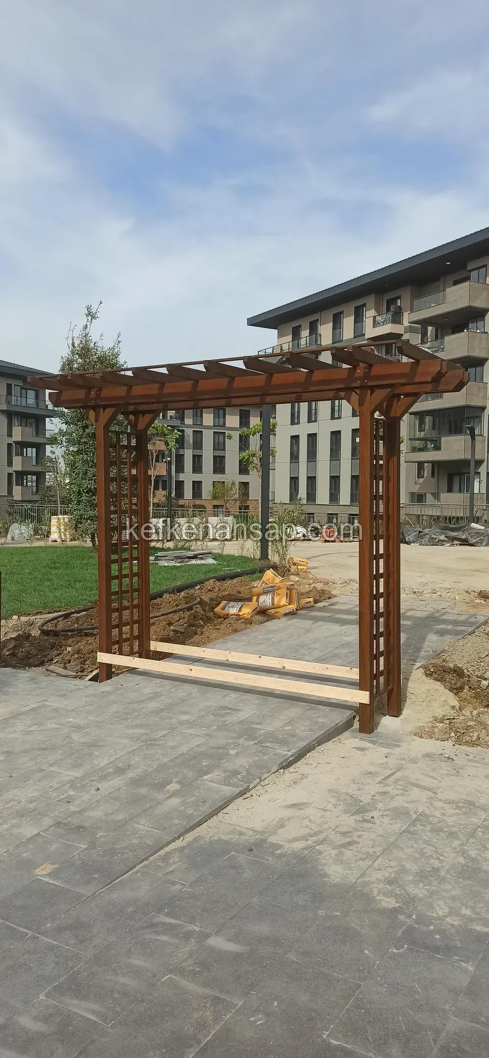 Taş döşeli yolun üzerinde kafes yanlı ahşap pergola kemeri, çimlik alan ve arkada apartman blokları