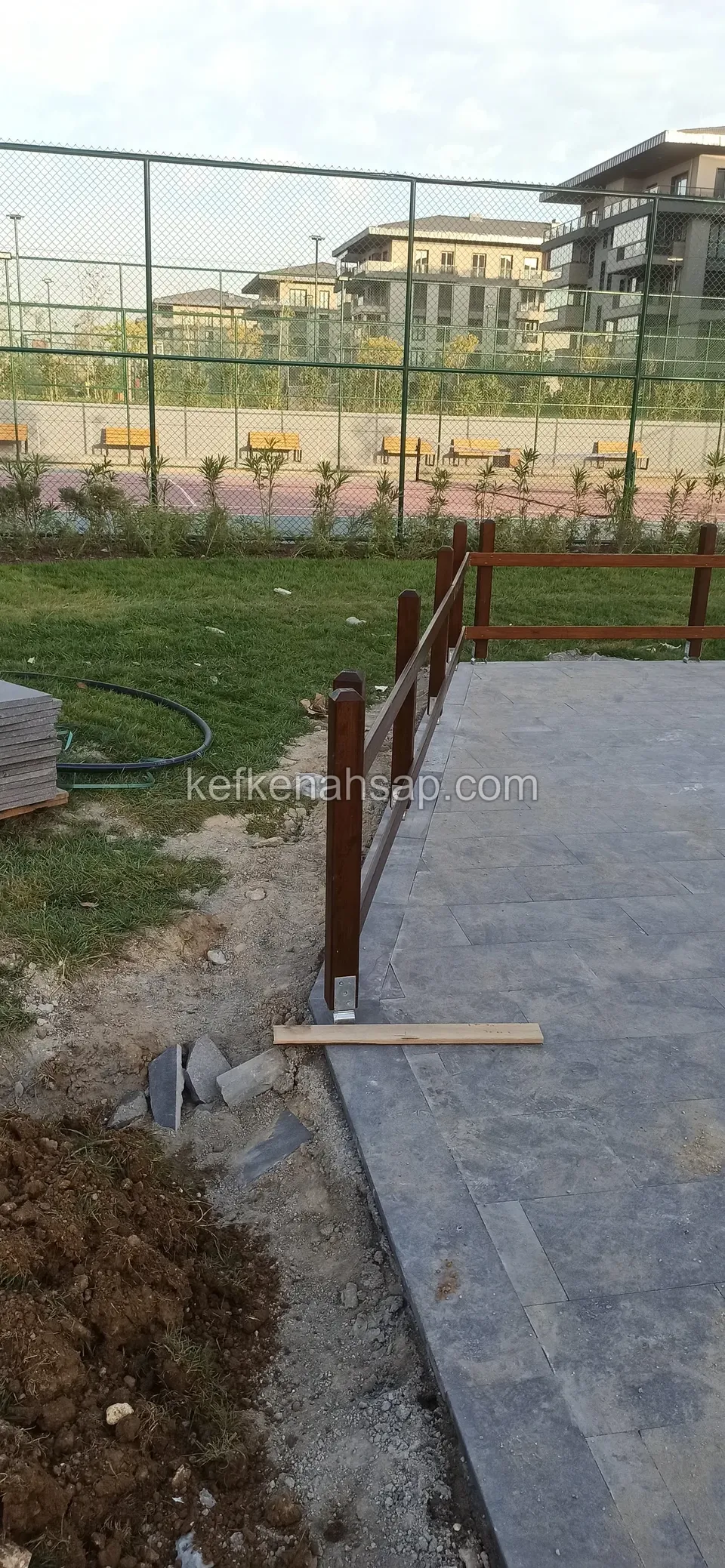 Dark timber fence posts set in the soil beside a grey stone-paved path, with a board and a pile of earth on the ground