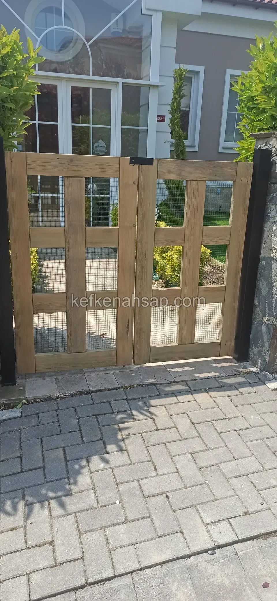 Kilitli taş döşeme yolun başında, kare gözlü kafes desenli çift kanatlı açık kahve ahşap bahçe kapısı