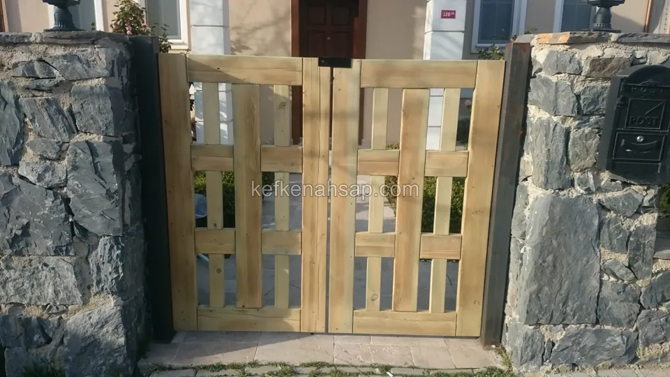 İki taş duvar arasında, bölmeli açık renk ahşap çift kanatlı kapı; sağdaki duvarda siyah posta kutusu, arkada ev