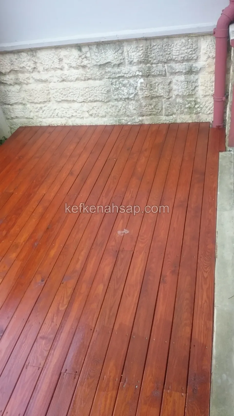 Taş kaplı bir duvarın dibinde, yağmur borusunun yanında kırmızı kahve tonlu dar deck alanı