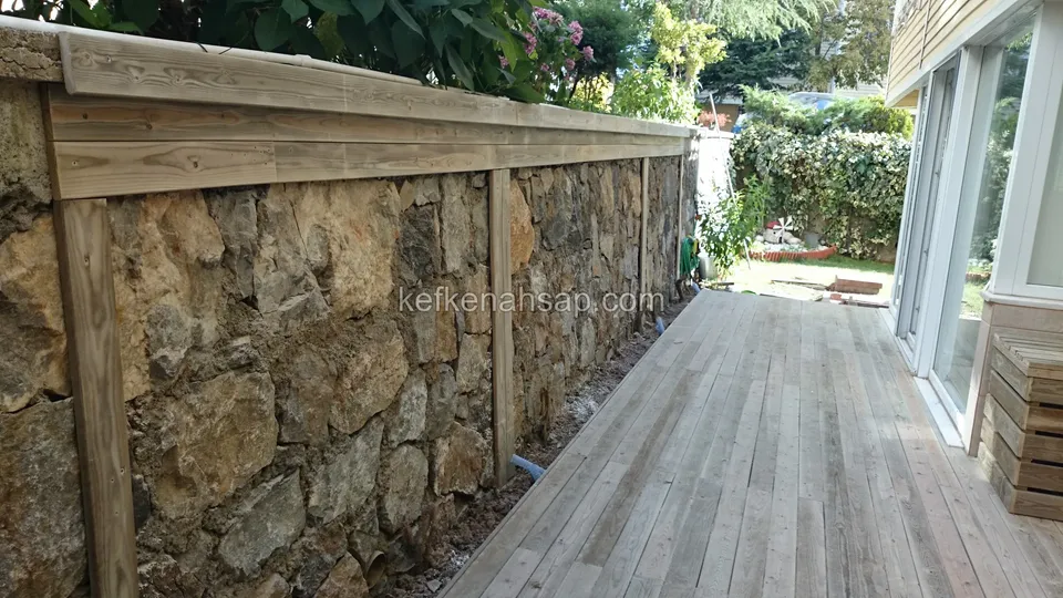 ديكينغ من الصنوبر المعالج بدرجة رمادية يمتد بين جدار حجري وباب زجاجي منزلق وتظهر خلفه خضرة الحديقة