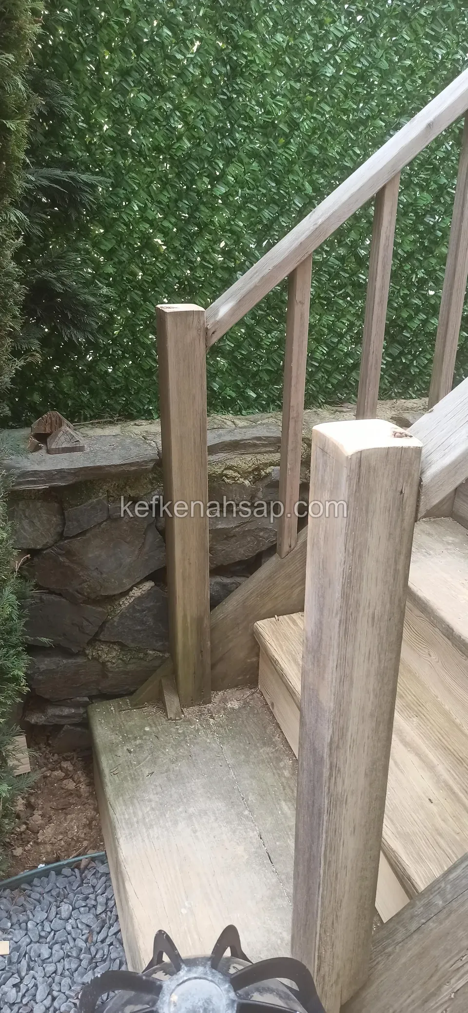 Holzstufen und Lattengeländer mit kräftigen Pfosten neben einer Stützmauer aus Stein, dahinter eine grüne Hecke