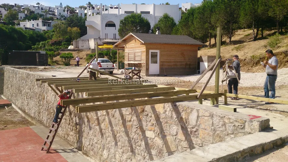 Taş duvarın üstünde pergola kirişlerini yerleştiren işçiler, arkada ahşap kabin ve beyaz evler
