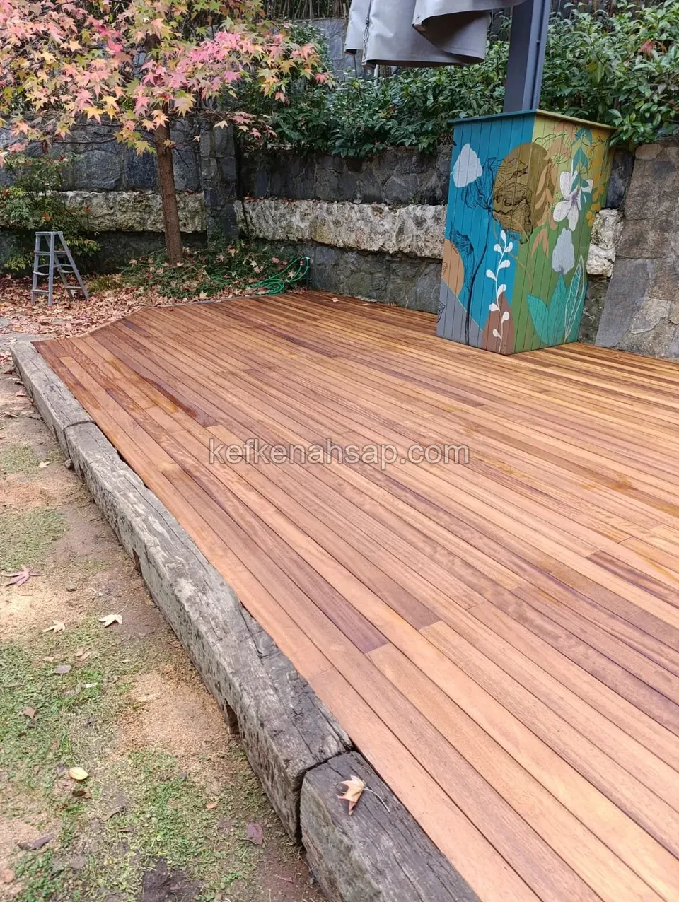 Honey-toned wooden garden deck edged with old timber beams, in front of a stone wall and a flower-painted post