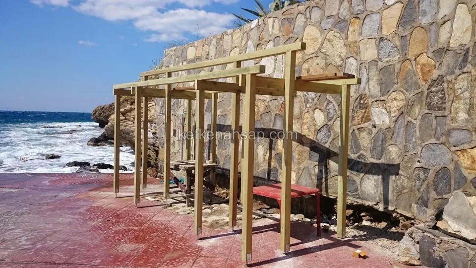 Taş duvarın önünde peş peşe dizili ahşap pergola iskeletleri, kırmızı bir bank ve kıyıya vuran dalgalar