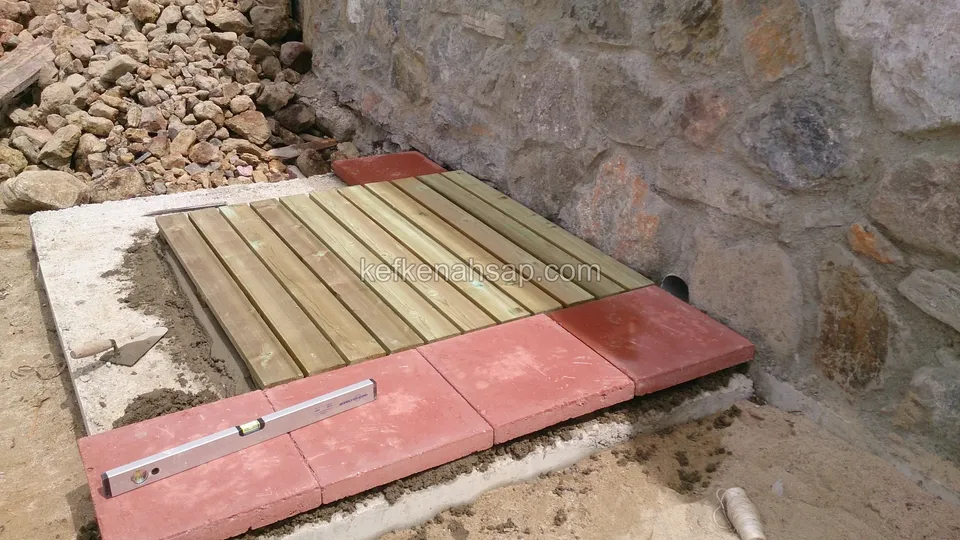 Taş duvarın önündeki beton zemine yerleştirilen küçük deck paneli; çevresinde kırmızı beton plakalar