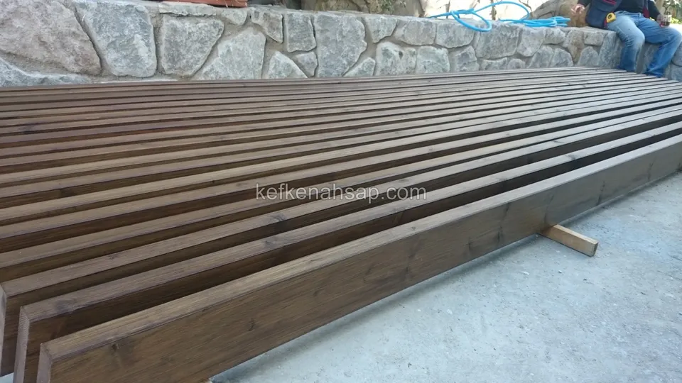 Beton zemin üzerinde taş istinat duvarına paralel dizilmiş koyu kahve tonlu ahşap kirişler